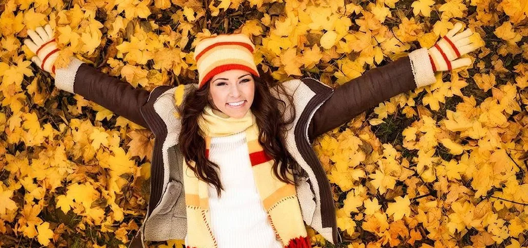 A woman in a brown jacket, striped hat and scarf lying among golden autumn leaves